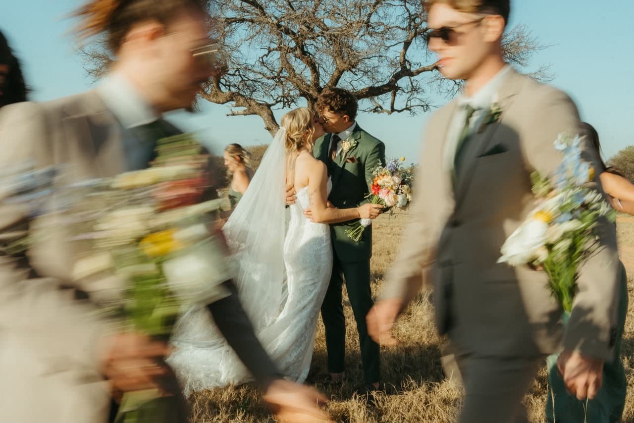 Emma and Juan Théron kissing under a thorn tree at their own wedding while their wedding party walks past